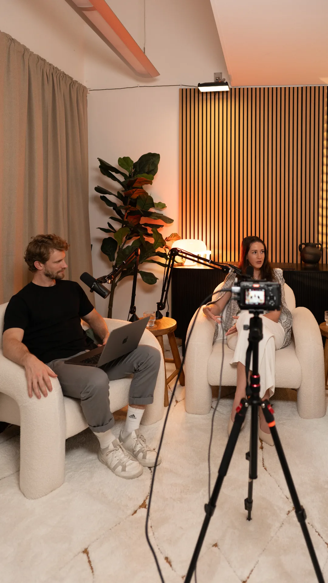 Two people seated in armchairs facing microphones on a warmly lit podcast set