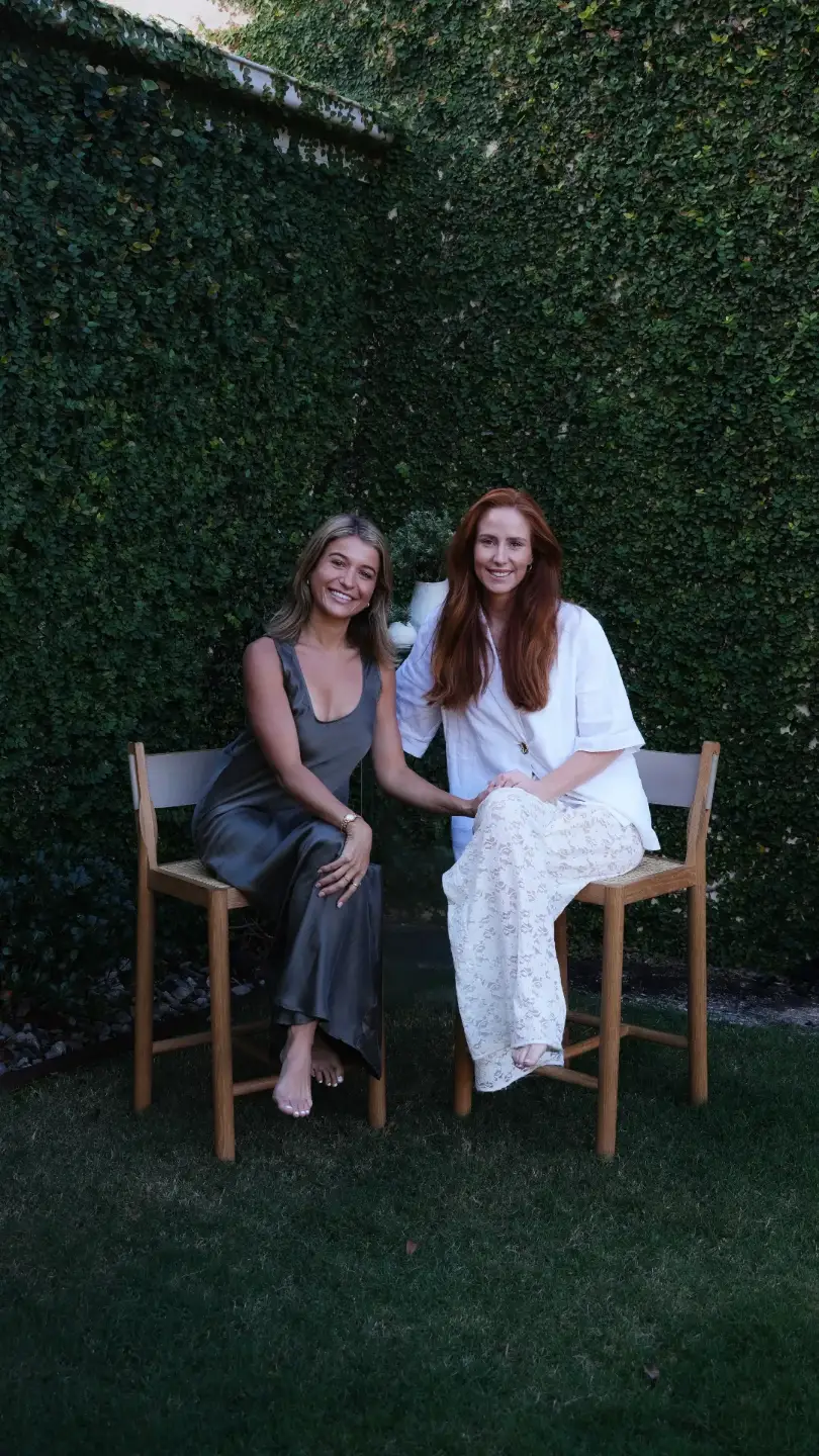 Two women seated on timber chairs in front of a dense green hedge